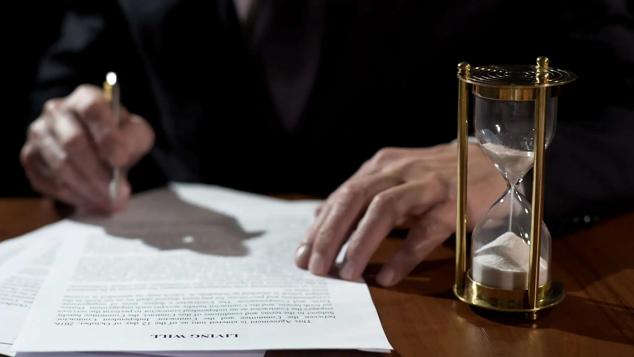 Person signing a document beside an hourglass on a wooden table.